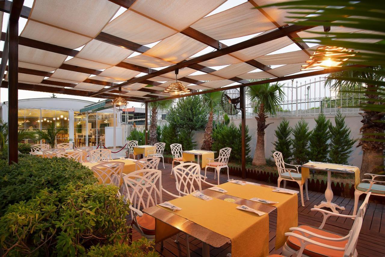 Sealife Family Resort Hotel Antalya Luaran gambar The photo depicts an outdoor dining area of a restaurant featuring a pergola with fabric coverings that provide shade. The space is arranged with several tables adorned with bright yellow tablecloths. The chairs are designed with a distinctive white 