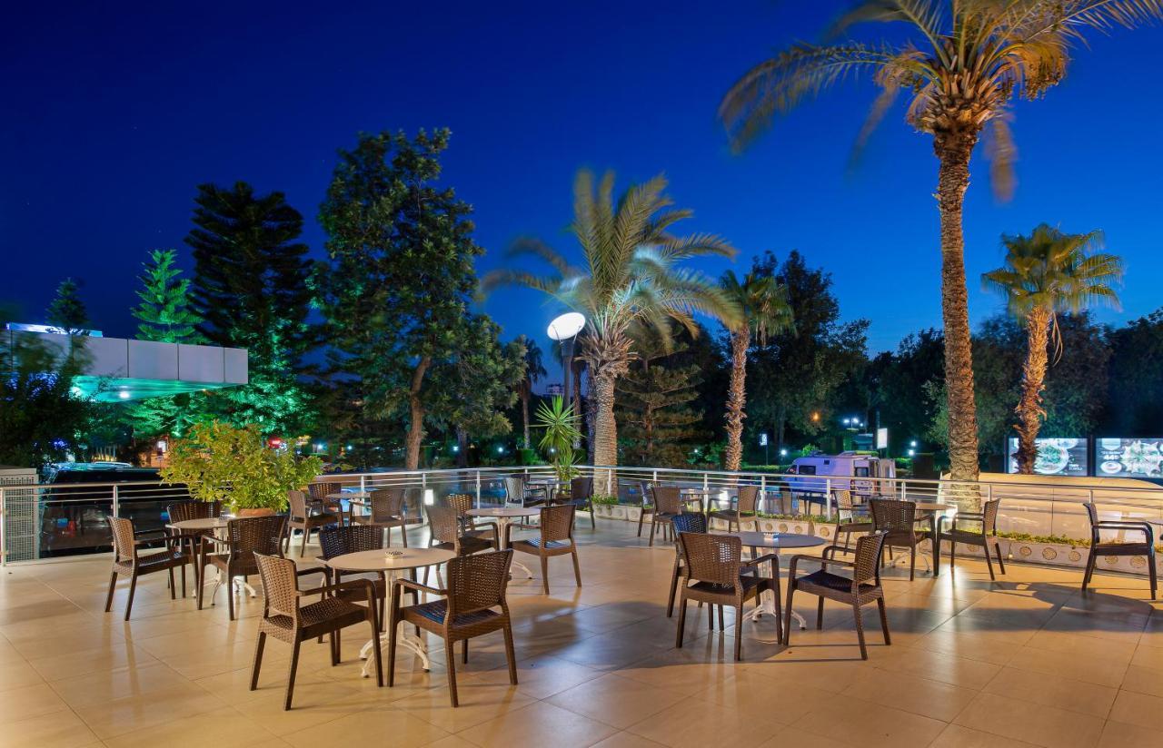 Sealife Family Resort Hotel Antalya Luaran gambar The photo depicts an outdoor dining area, likely part of a restaurant or café, set in the evening. There are several tables with chairs arranged across a tiled patio. The space is surrounded by tall palm trees and greenery, creating a tropical ambian
