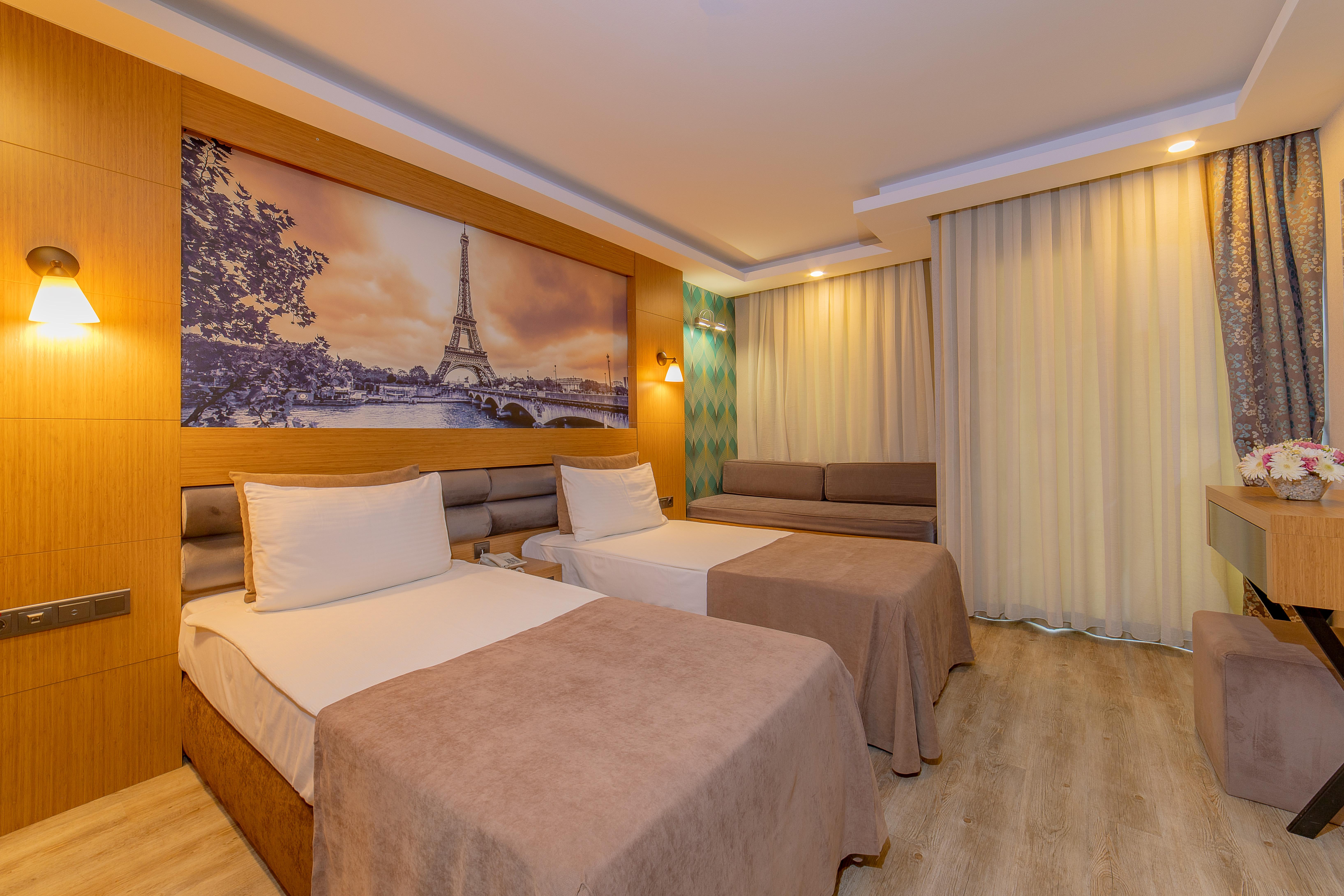 Sealife Family Resort Hotel Antalya Luaran gambar The photo shows a modern hotel room featuring two beds with beige bedspreads. There is a large wall mural depicting a scenic view of the Eiffel Tower in Paris. The room is well-lit, with soft lighting fixtures on the walls. To the right, there is a s
