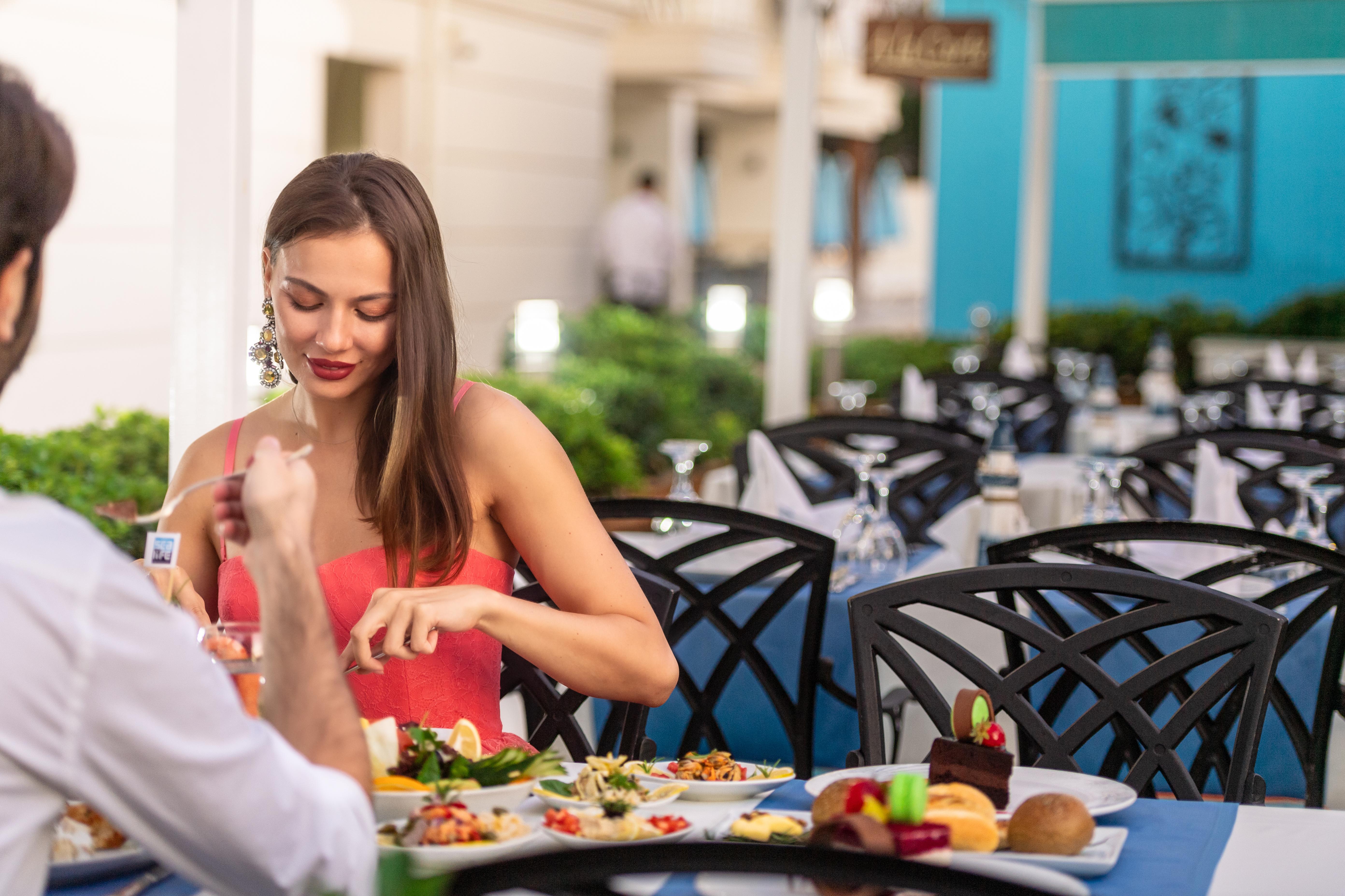 Sealife Family Resort Hotel Antalya Luaran gambar The photo shows a dining scene at a restaurant. A woman in a red dress is seated at a table, interacting with her meal. She appears to be taking time to enjoy her food. The table is set with various dishes, including salads and desserts. The backgrou