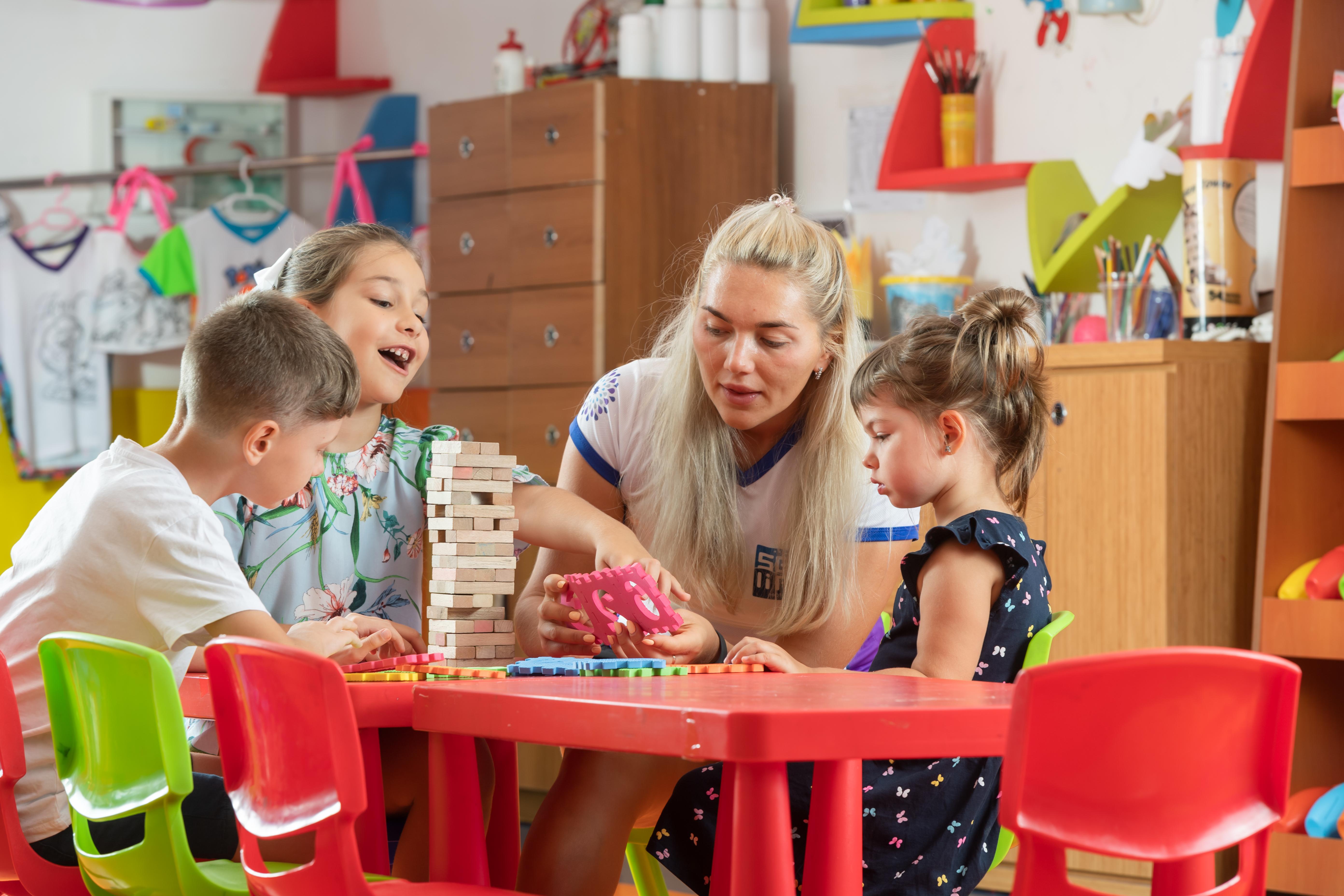 Sealife Family Resort Hotel Antalya Luaran gambar The photo depicts a playful and engaging scene in a classroom or activity space. There are four children sitting at a bright red table, focused on a game. Two girls, one wearing a colorful floral shirt and the other in a dark dress with ruffles, are 