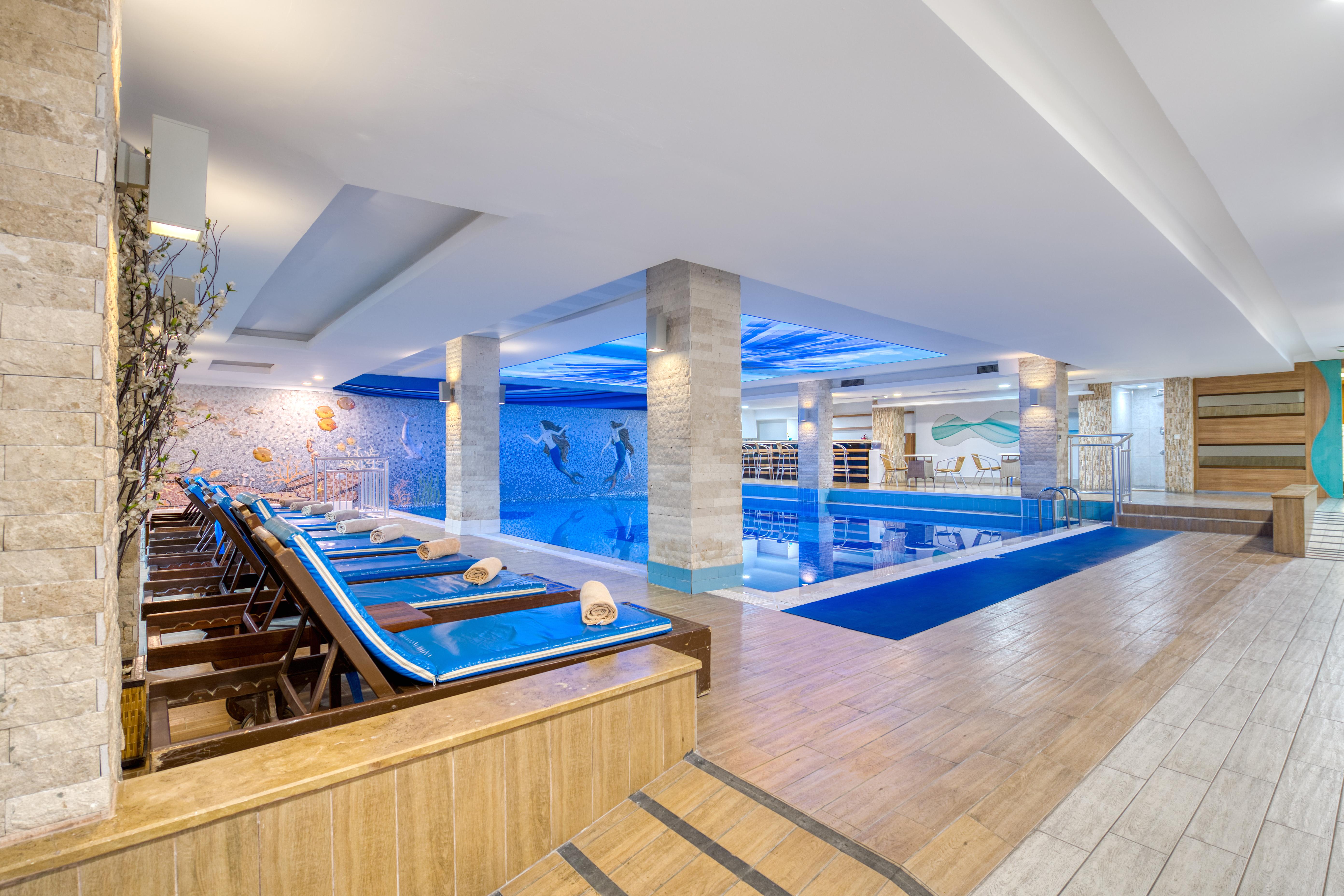 Sealife Family Resort Hotel Antalya Luaran gambar The image shows an indoor swimming pool area. The pool is illuminated with soft blue lighting, creating a relaxing atmosphere. Alongside the pool, there are several lounge chairs arranged, some of which have rolled towels placed on them. The flooring