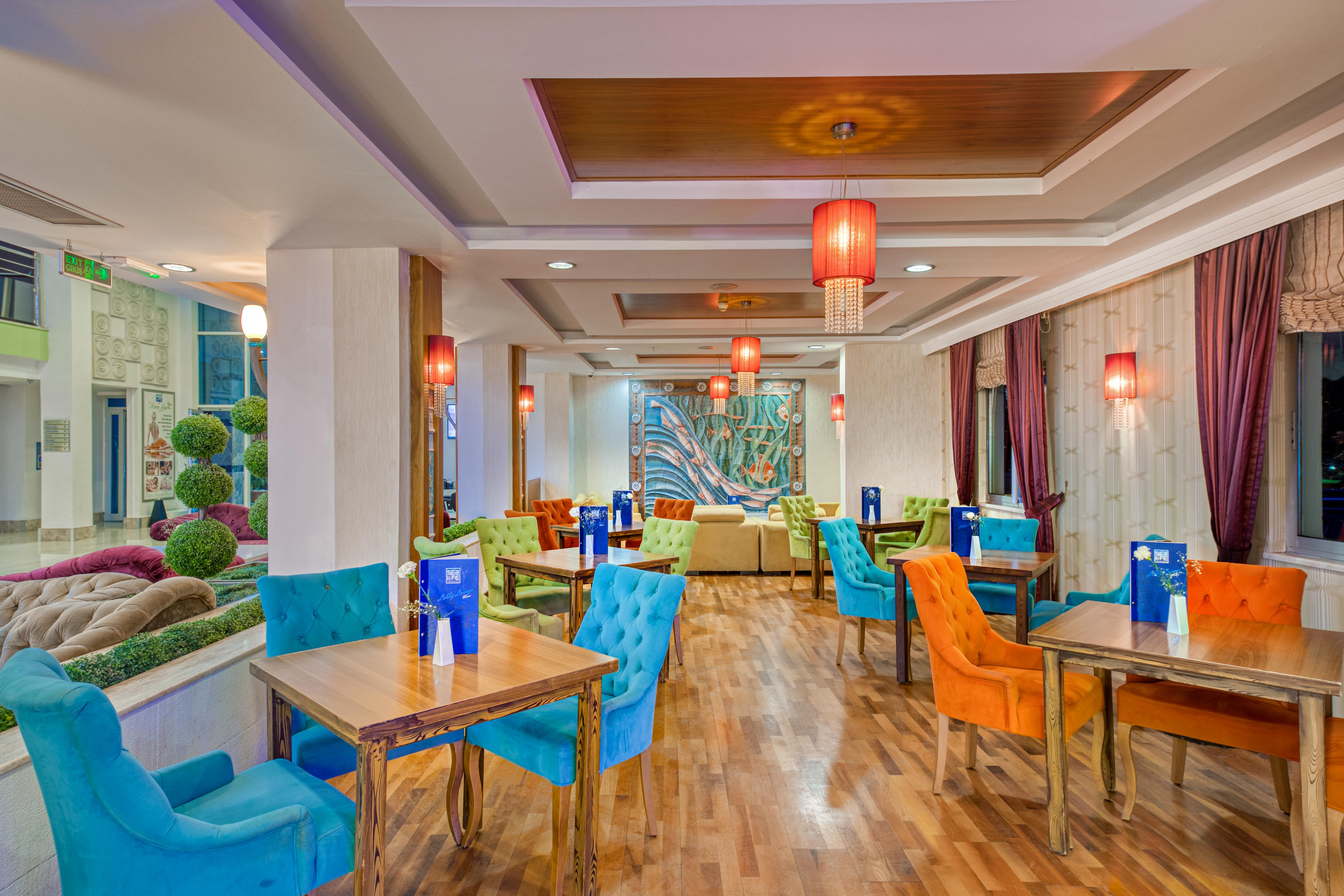 Sealife Family Resort Hotel Antalya Luaran gambar The photo shows a stylish dining area with a contemporary design. The space features a combination of colorful chairs in blue and orange, and wooden tables arranged throughout. The walls have a warm, neutral tone and are complemented by decorative el