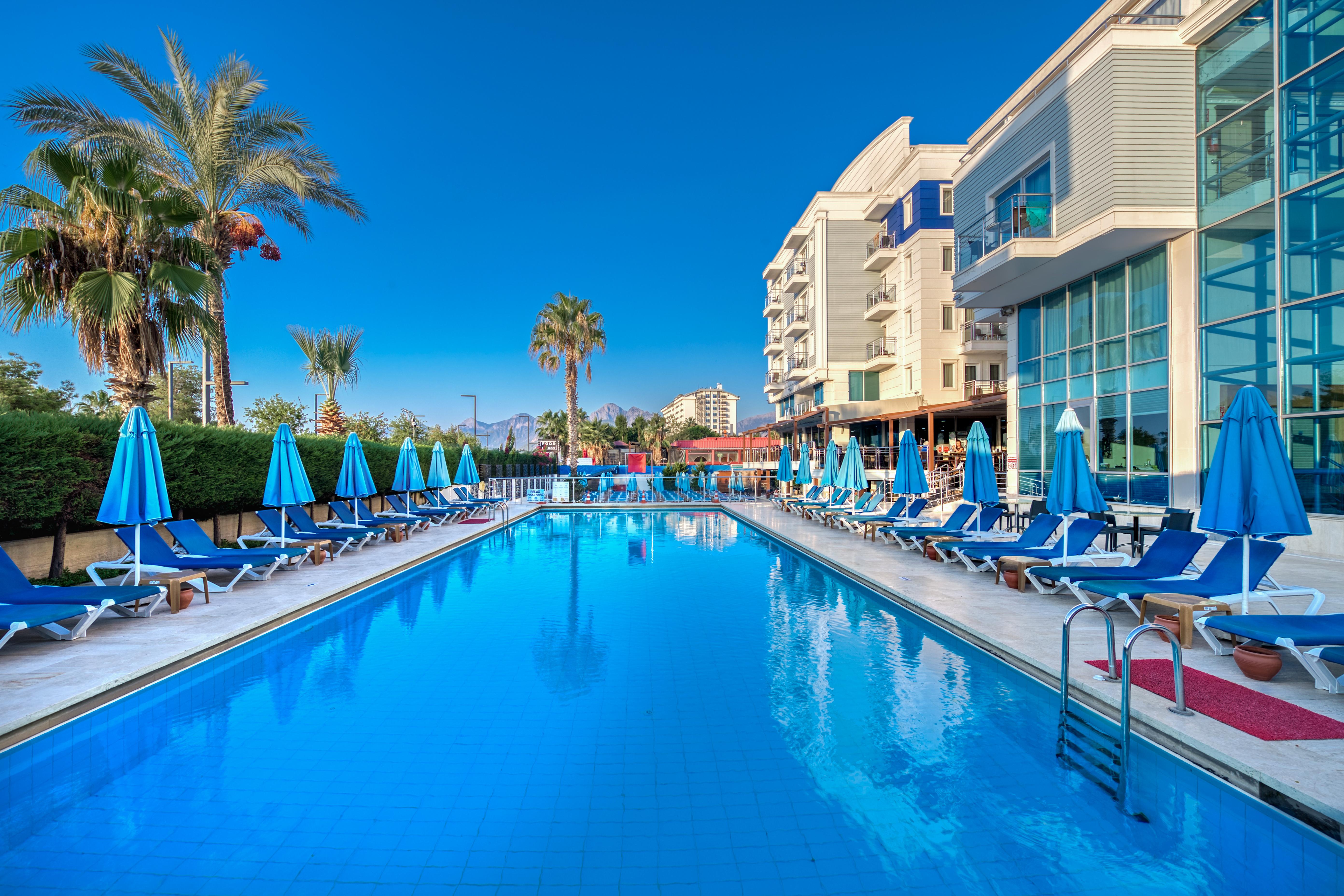 Sealife Family Resort Hotel Antalya Luaran gambar The photo depicts a serene poolside scene at a hotel or resort. In the foreground, a large blue swimming pool reflects the clear sky, with comfortable lounge chairs arranged neatly around it under blue umbrellas. Lush palm trees add to the tropical a