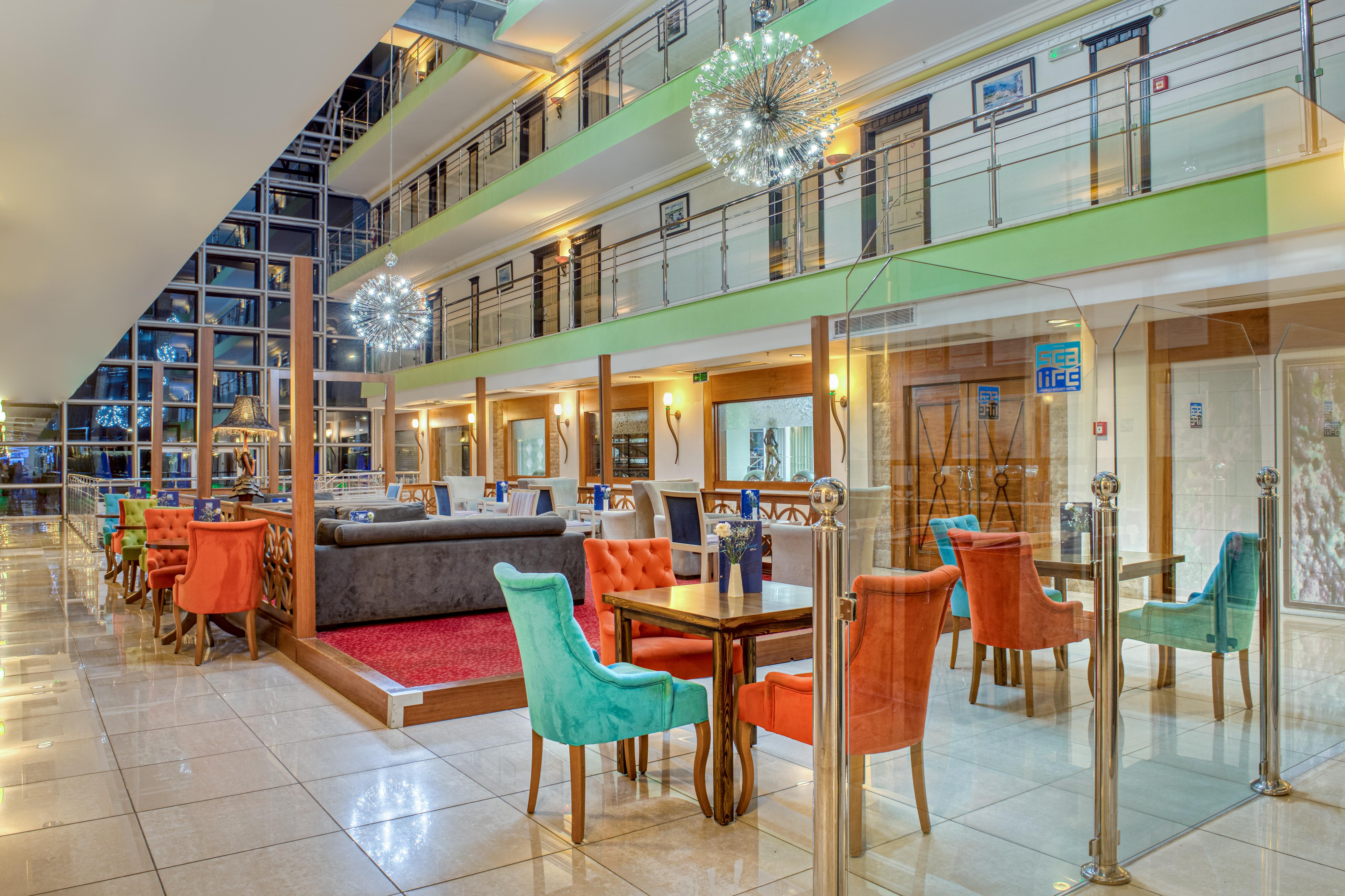Sealife Family Resort Hotel Antalya Luaran gambar The photo shows the interior of a modern hotel lobby or dining area. It features a spacious room with multiple levels visible. The decor includes colorful chairs in orange and turquoise, along with wooden tables. 

There are glass barriers separating