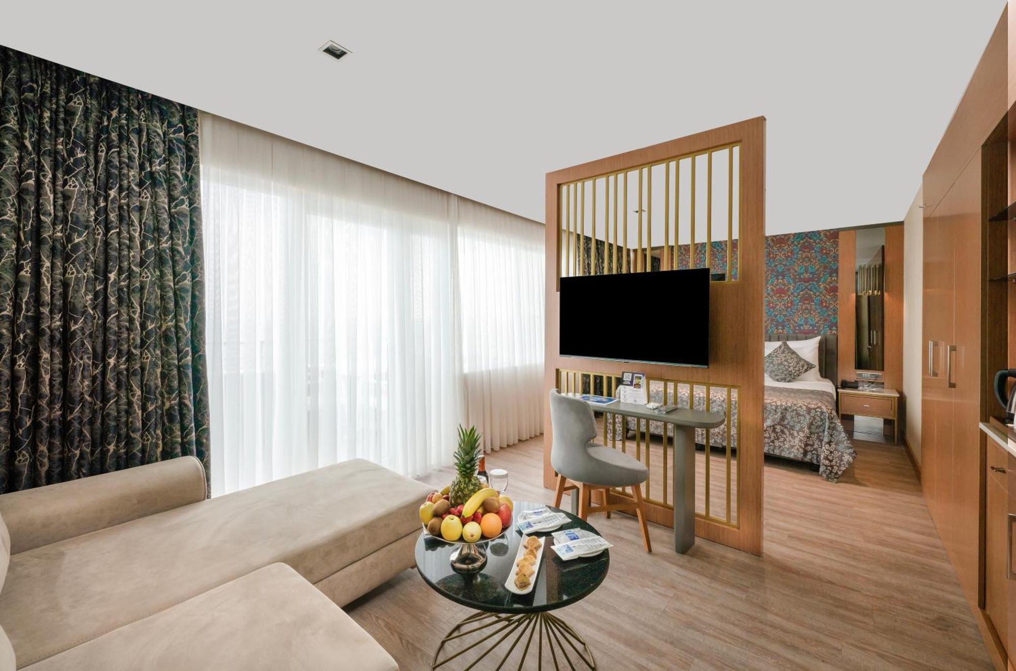 Sealife Family Resort Hotel Antalya Bilik gambar The photo shows a modern hotel room that features a stylish and functional design. On one side, there is a comfortable light-colored sofa positioned next to a round coffee table adorned with a variety of fruits and snacks. The coffee table has a slee